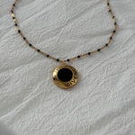 Reworked vintage Givenchy pendant necklace featuring a round gold medallion with black enamel center and GIVENCHY script detailing, suspended on a delicate gold chain with black beaded accents, laid flat on a soft white fabric background.