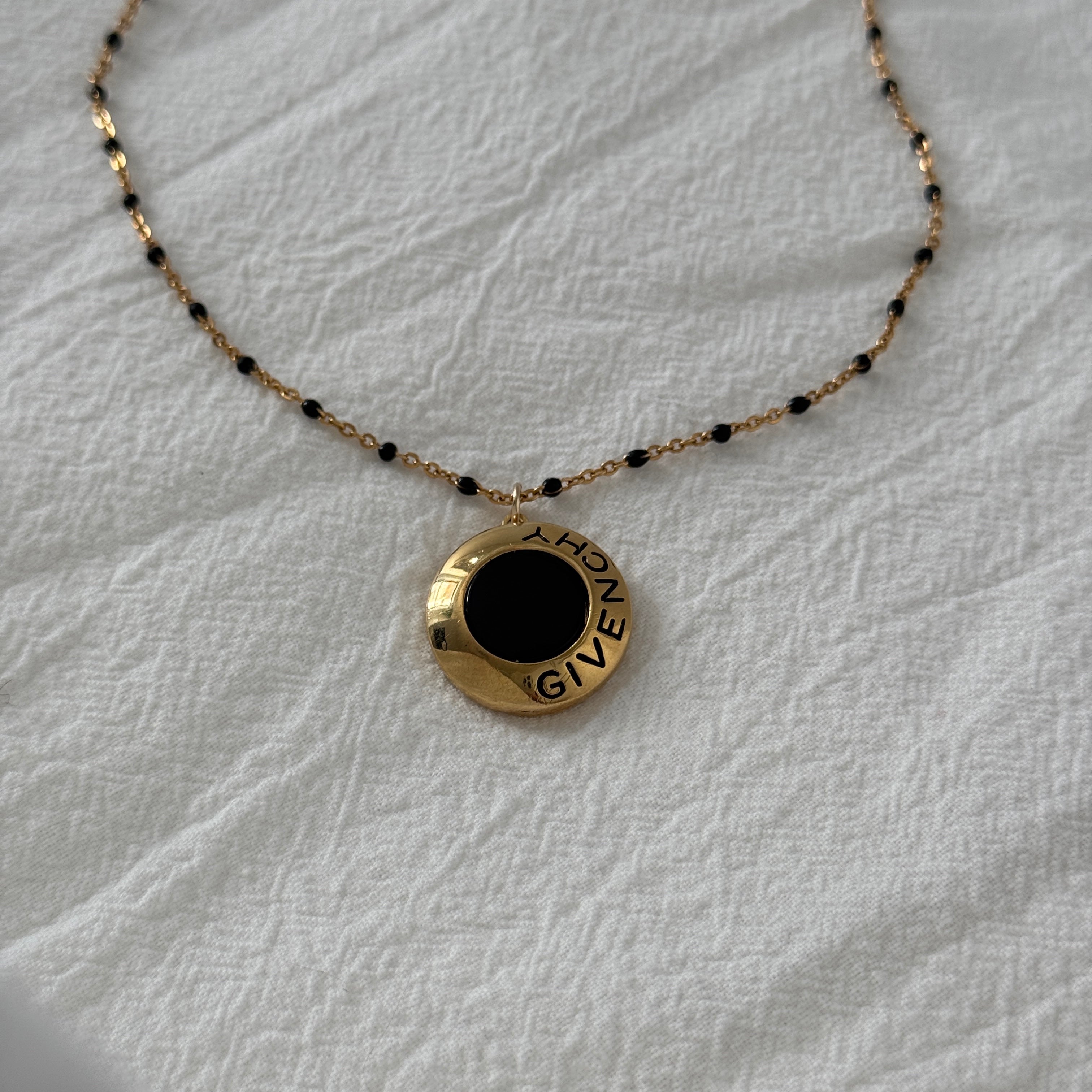 Reworked vintage Givenchy pendant necklace featuring a round gold medallion with black enamel center and GIVENCHY script detailing, suspended on a delicate gold chain with black beaded accents, laid flat on a soft white fabric background.