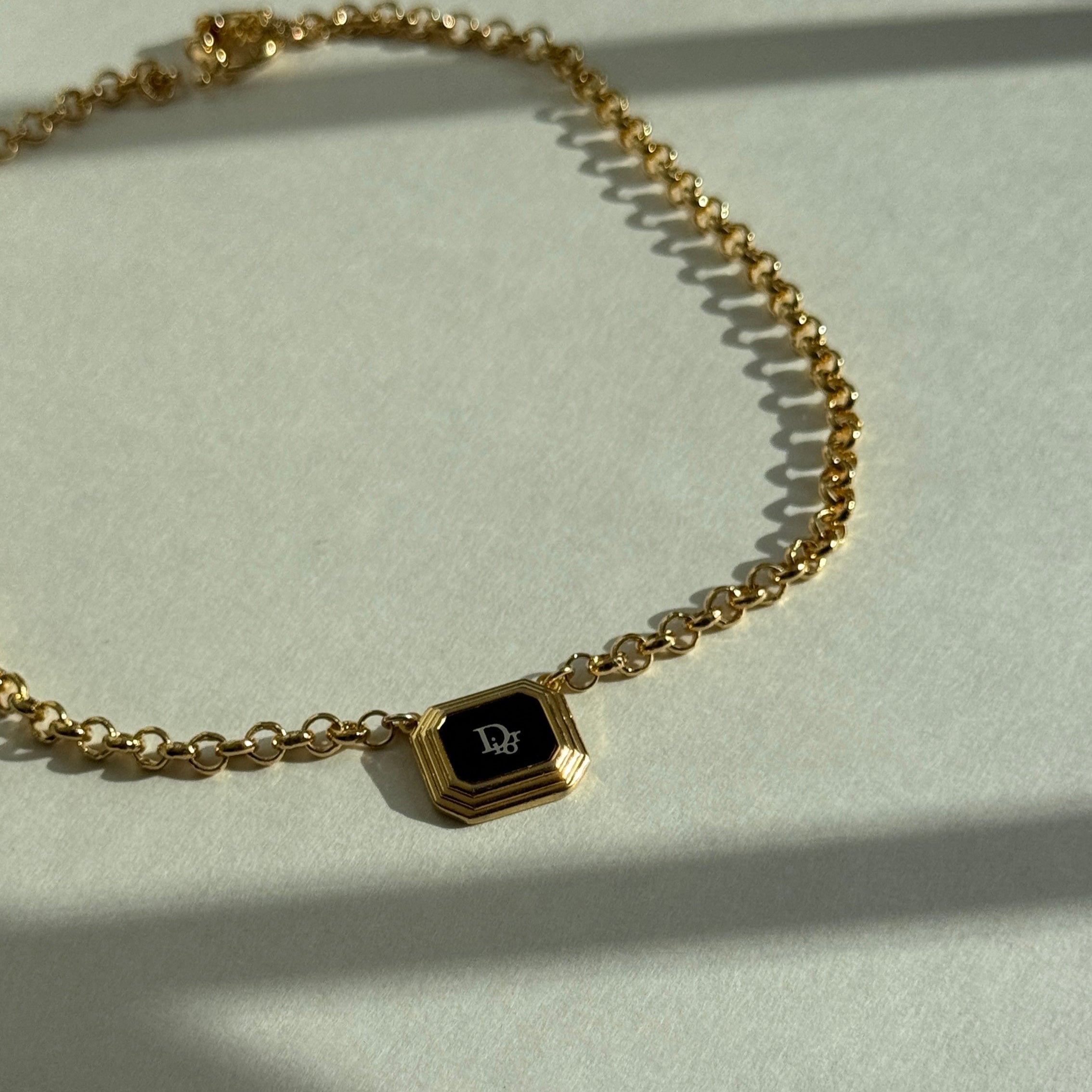 Reworked vintage Dior pendant necklace featuring an octagonal gold frame with black enamel center and white Dior monogram, suspended on a gold rolo chain, styled flat on a soft white fabric background
