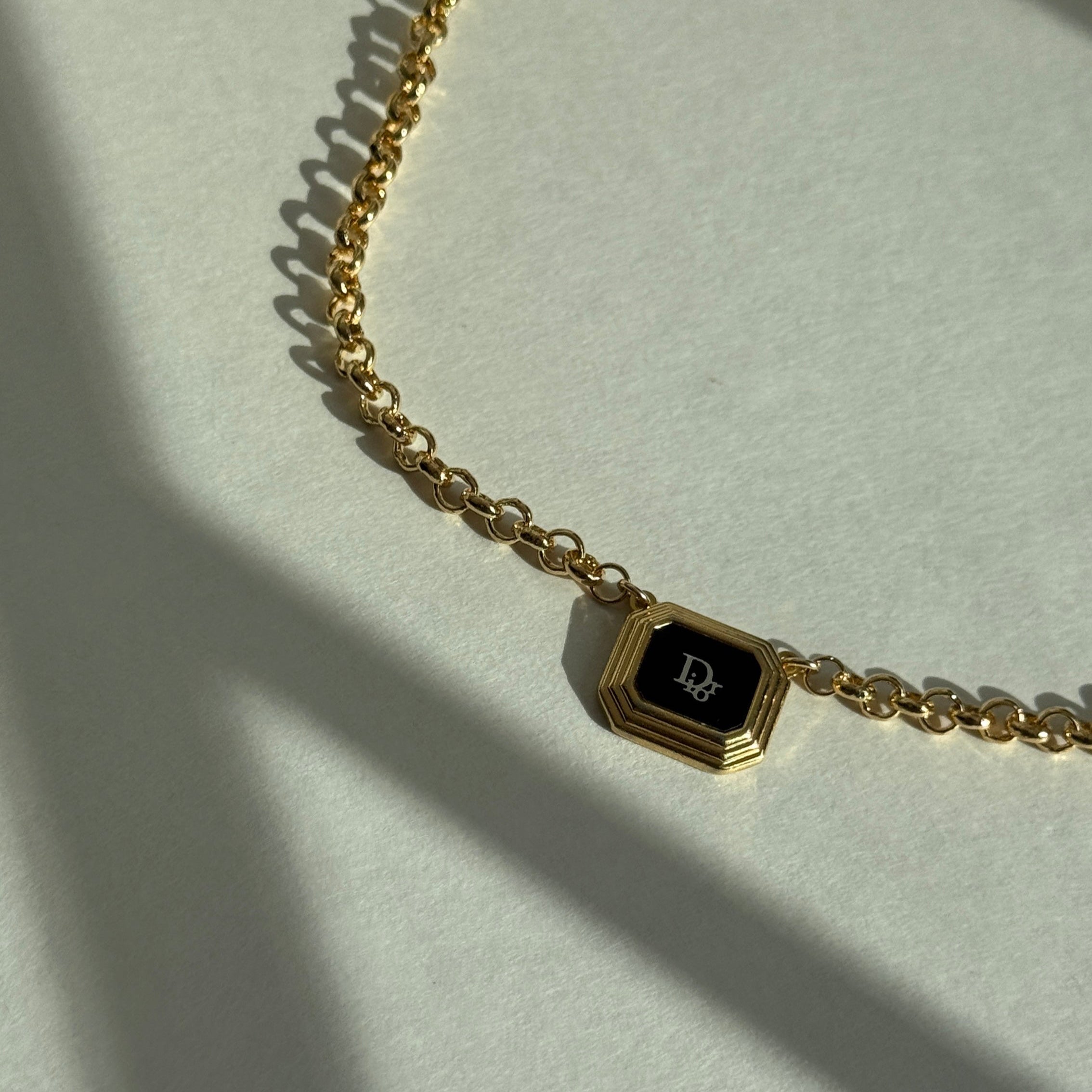 Reworked vintage Dior pendant necklace featuring an octagonal gold frame with black enamel center and white Dior monogram, suspended on a gold rolo chain, styled flat on a soft white fabric background