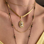 A close-up of a person wearing a layered gold necklace set with a central oval pendant featuring a white pearl-like center and dark green bead accents.