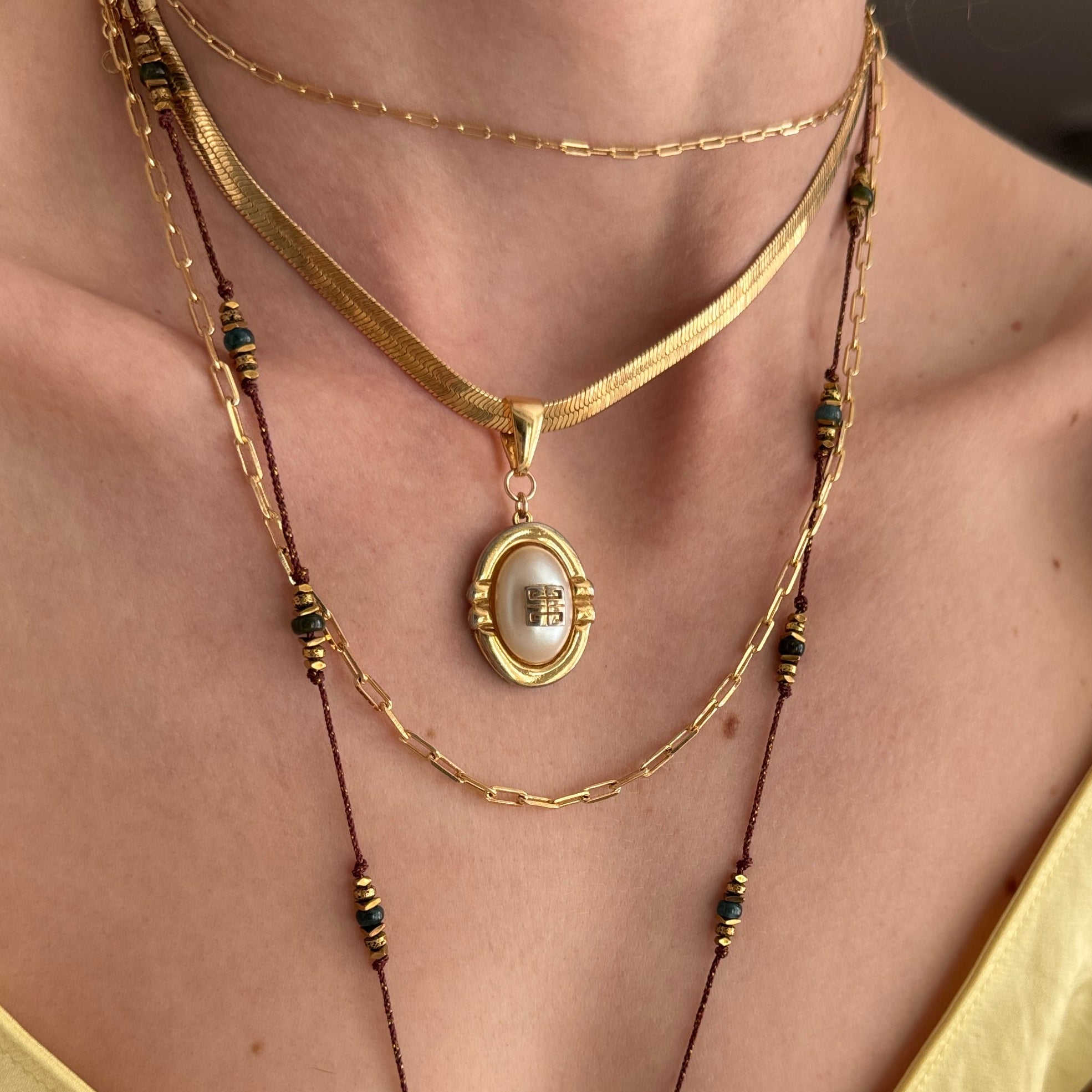 A close-up of a person wearing a layered gold necklace set with a central oval pendant featuring a white pearl-like center and dark green bead accents.