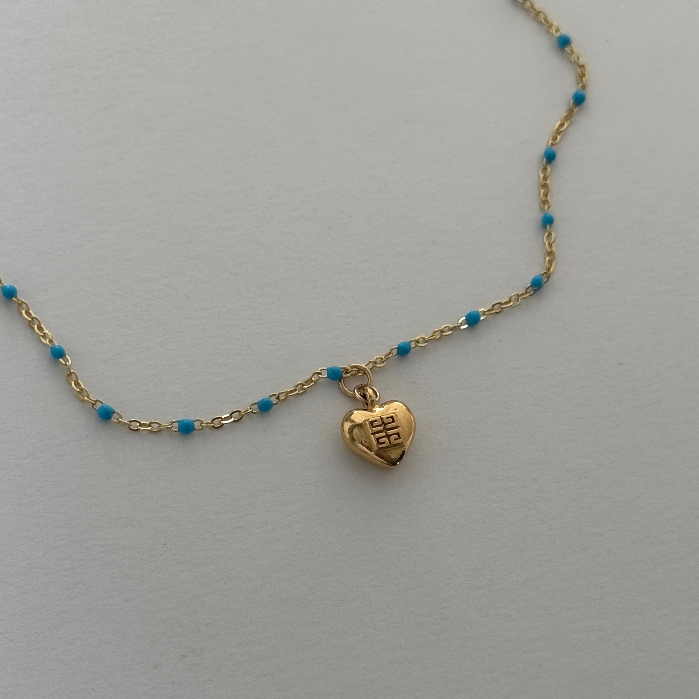 Close-up of a vintage mini Givenchy heart charm necklace, featuring an authentic gold-toned heart pendant on a delicate enamel colored blue chain. A rare and timeless piece, repurposed for modern wear, blending classic elegance with effortless everyday luxury