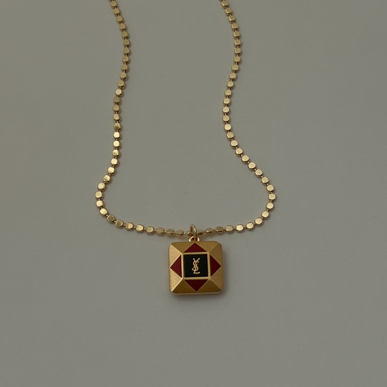 A vintage square necklace with a gold chain and a geometric pendant featuring a red and yellow YSL logo.