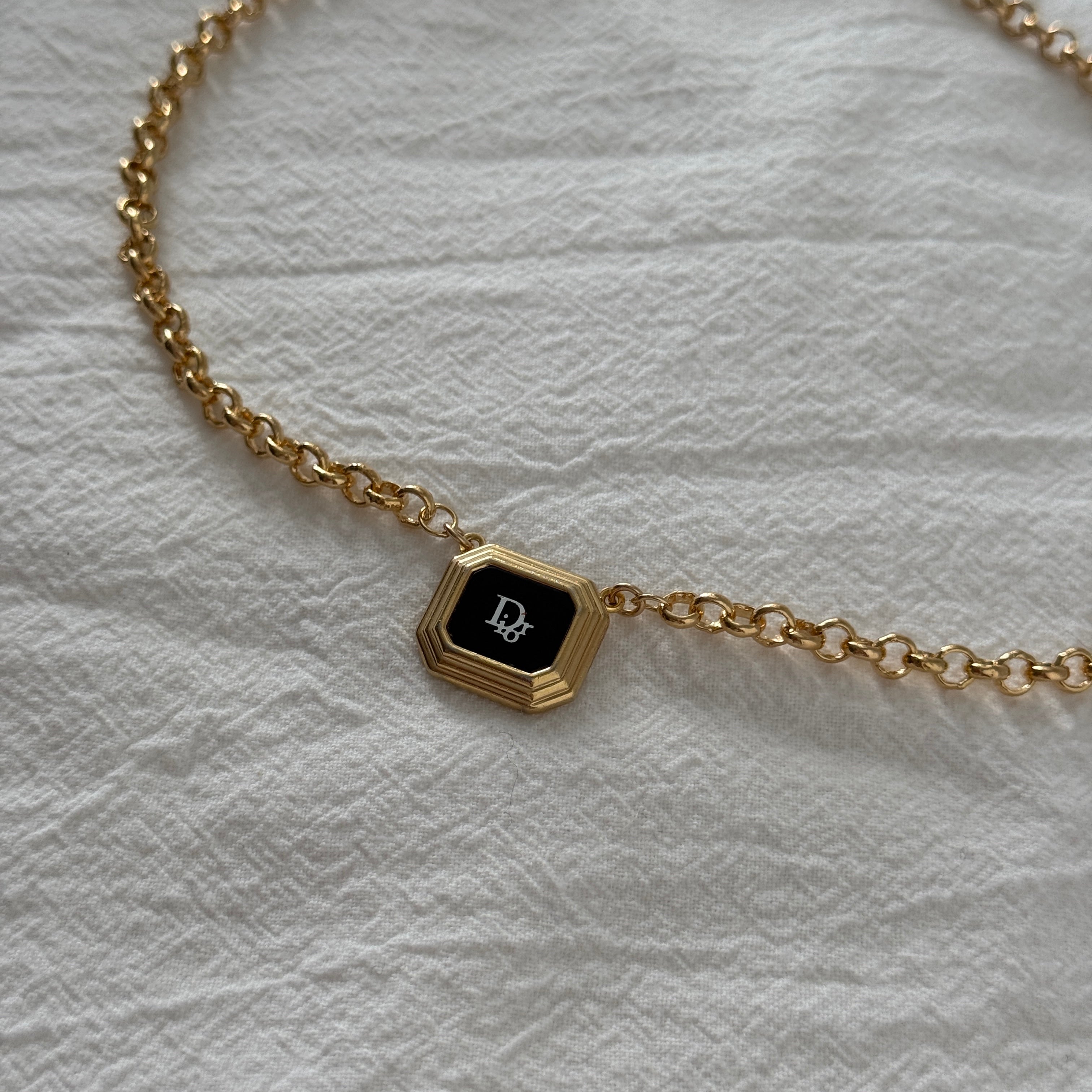 Reworked vintage Dior pendant necklace featuring an octagonal gold frame with black enamel center and white Dior monogram, suspended on a gold rolo chain, styled flat on a soft white fabric background