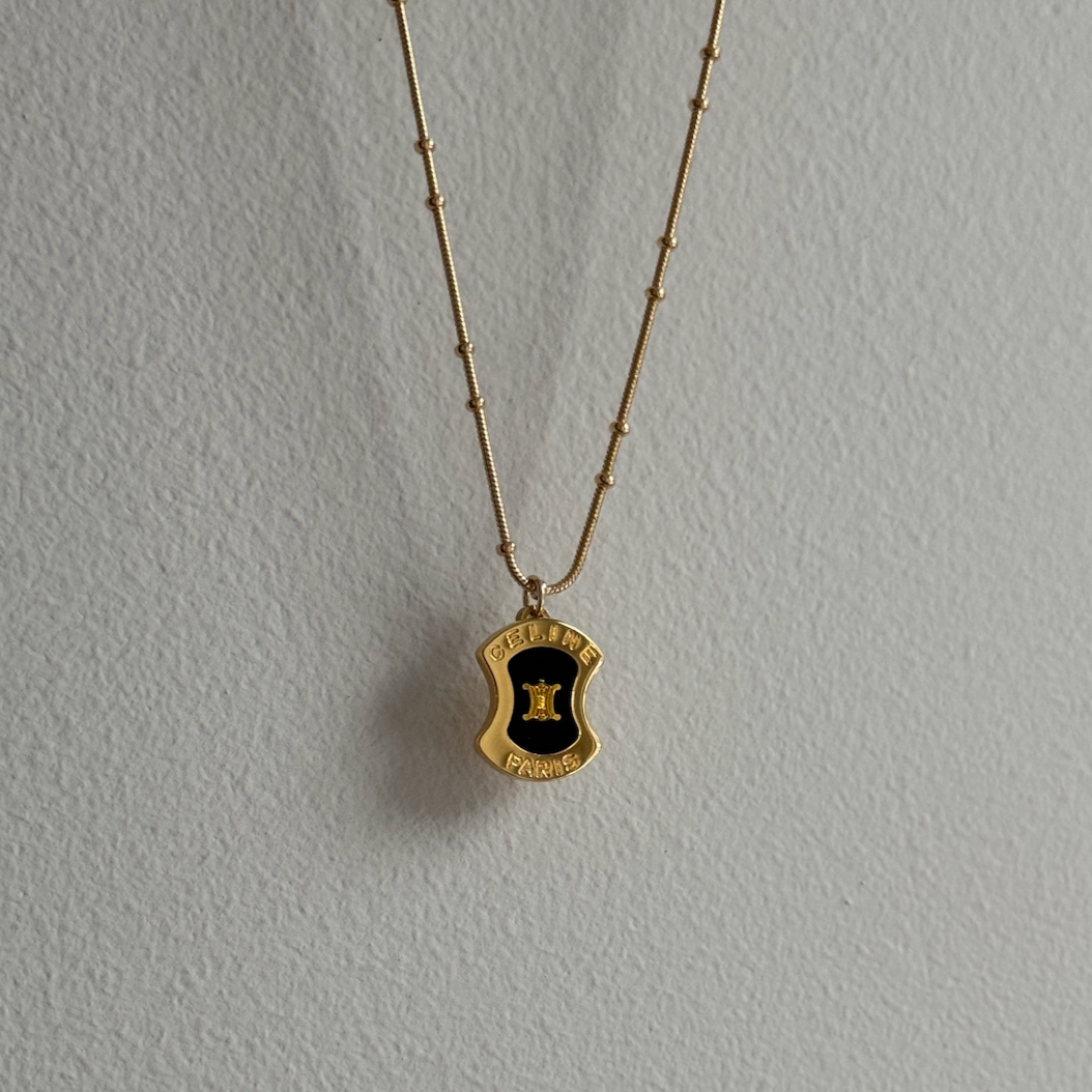 Reworked vintage Celine mini plaque pendant featuring a black and gold enamel crest marked 'CELINE PARIS,' suspended on a gold textured bar and satellite chain, styled against a soft white fabric background